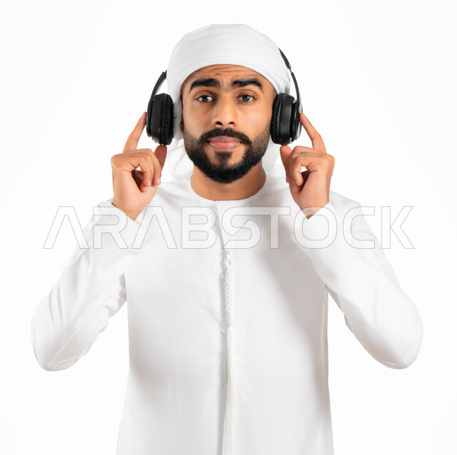 سماع الأغاني والموسيقى والصوتيات، بورتريه لرجل عربي خليجي إماراتي يرتدي العمامة والكندورة الإماراتية وسماعات الرأس الصوتية، التسلية والترفيه والإستمتاع بإستخدام تقنية الهيدفون، خلفية بيضاء