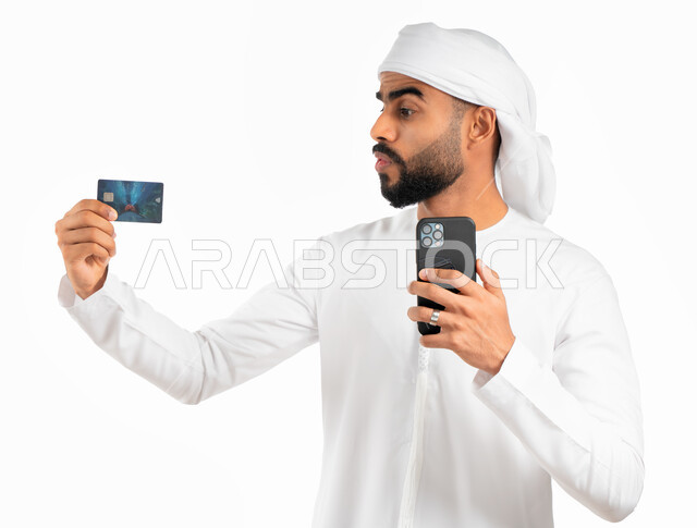 الشراء والدفع من خلال برامج وتطبيقات الجوال، بورتريه لرجل عربي خليجي إماراتي يرتدي الكندورة والعمامة الإماراتية ويمسك بيده الهاتف المحمول وبطاقة الإئتمان البنكية، إمكانية تسديد الفواتير بالبطاقة الإلكترونية، خلفية بيضاء