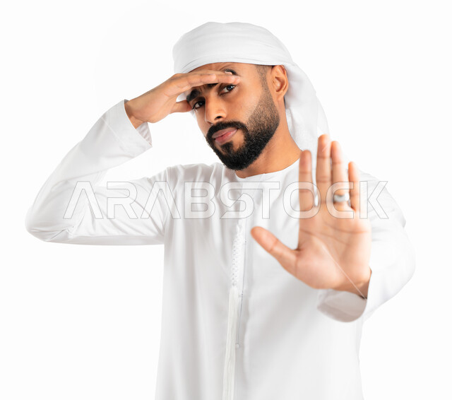 Request to stop and refrain from doing something, non-acceptance and refusal to harm, a portrait of an Arab Emirati man wearing a kandura and an Emirati turban and placing his hand on his head and raising his other hand up in a stop sign, gestures of fear