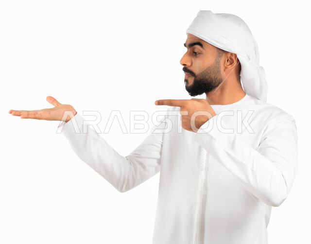 إيماءات حمل ورفع شيء ما، بورتريه لرجل عربي خليجي إماراتي يرتدي العمامة والكندورة الإماراتية البيضاء ويفتح يد ويشير باصبع السبابة نحو اليد الاخرى، الإشارة نحو هدف ما، حركة أصبع وإشارات، خلفية بيضاء