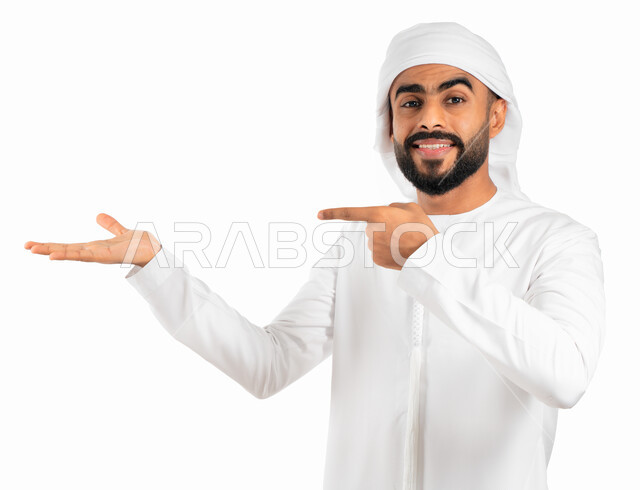 الإشارة نحو هدف ما، حركة أصبع وإشارات، بورتريه لرجل عربي خليجي إماراتي مبتسم يرتدي العمامة والكندورة الإماراتية البيضاء ويفتح يد ويشير باصبع السبابة نحو اليد الاخرى، إيماءات حمل ورفع شيء ما، خلفية بيضاء