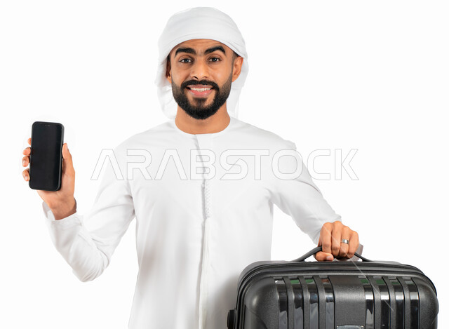 عرض مواقع وتطبيقات السياحة والسفر من خلال الجوال، بورتريه لرجل عربي خليجي إماراتي يرتدي الكندورة والعمامة الإماراتية ويمسك بيده الهاتف المحمول بشاشة سوداء فارغة وحقيبة السفر، الاستعداد للسفر والسياحة وقضاء إجازة صيفية ممتعة، خلفية بيضاء