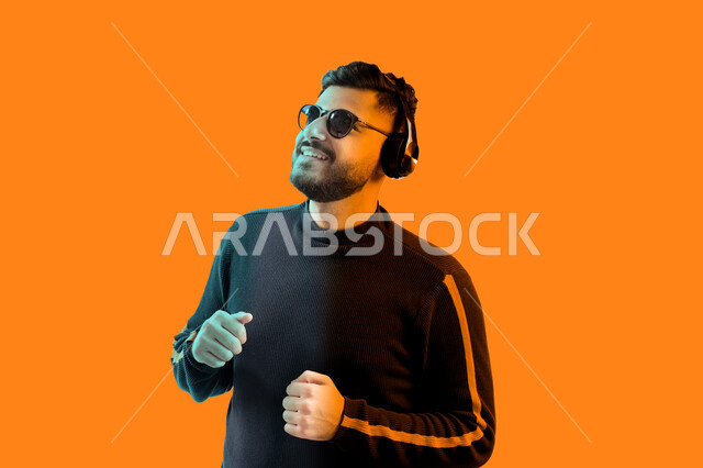 إيماءات و تعابير وجه ويدين تدل على الاندماج مع الأغنية، الاستماع إلى الأغاني بواسطة الهيدفون، بورتريه مقرب لشاب سعودي يرتدي النظارات الشمسية وسماعات رأس في استديو صوتي، رجل عربي إماراتي خليجي مبتسم يرتدي الزي الكاجوال ينظر إلى شيء ما، خلفية ملونة