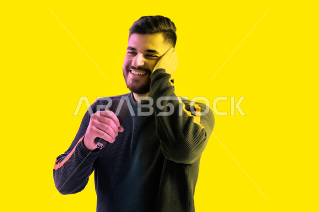 ايماءات تدل على الاندماج والتفاعل مع الغناء، الاستمتاع بأداء المواهب الصوتية، بورتريه مقرب لرجل عربي اماراتي خليجي يرتدي الزي الكاجوال يمسك بيده المايكروفون و يغني بحماس، شاب سعودي يقوم بممارسة موهبة الغناء، تسجيل مقطع صوتي غنائي، خلفية صفراء