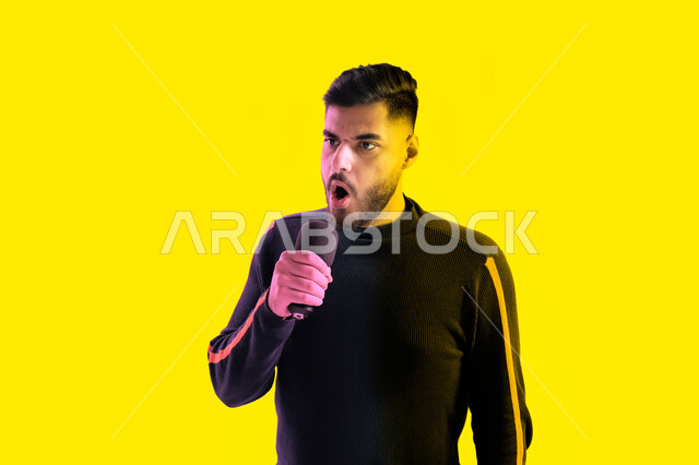 التدريب الصوتي في الاستديو، بورتريه مقرب لرجل عربي اماراتي خليجي يرتدي الزي الكاجوال يمسك بيده المايكروفون و يغني بحماس، ايماءات تدل على الاندماج والتفاعل مع الغناء، شاب سعودي يقوم بممارسة موهبة الغناء، تسجيل مقطع صوتي غنائي، خلفية صفراء