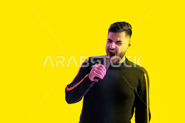 تقديم فقرات غنائية ترفيهية، بورتريه مقرب لرجل عربي اماراتي خليجي يرتدي الزي الكاجوال يمسك بيده المايكروفون و يغني بحماس، ايماءات تدل على الاندماج والتفاعل مع الغناء، شاب سعودي يقوم بممارسة موهبة الغناء، تسجيل مقطع صوتي غنائي، خلفية صفراء