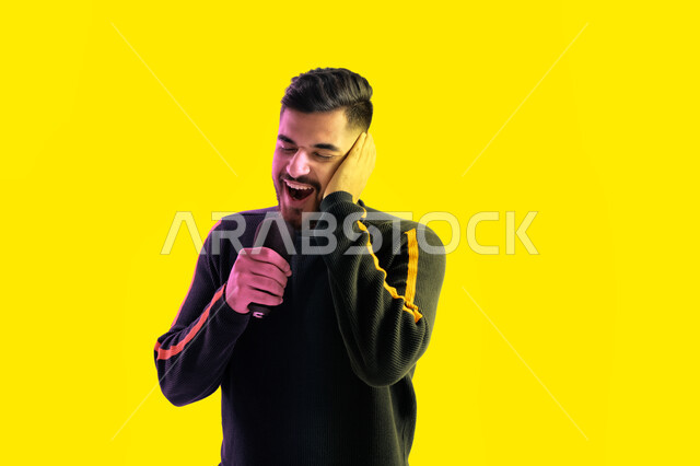  تسجيل مقطع صوتي غنائي، الاستمتاع بأداء المواهب الصوتية، بورتريه مقرب لرجل عربي اماراتي خليجي يرتدي الزي الكاجوال يمسك بيده المايكروفون و يغني بحماس، ايماءات تدل على الاندماج والتفاعل مع الغناء، شاب سعودي يقوم بممارسة موهبة الغناء، خلفية صفراء