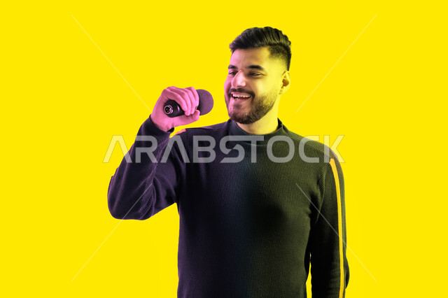 تسجيل مقطع صوتي غنائي، الاستمتاع بأداء المواهب الصوتية، بورتريه مقرب لرجل عربي اماراتي خليجي يرتدي الزي الكاجوال يمسك بيده المايكروفون و يغني بحماس، ايماءات تدل على الاندماج والتفاعل مع الغناء، شاب سعودي يقوم بممارسة موهبة الغناء، خلفية صفراء