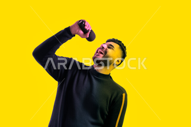تسجيل مقطع صوتي غنائي، الاستمتاع بأداء المواهب الصوتية، بورتريه مقرب لرجل عربي اماراتي خليجي يرتدي الزي الكاجوال يمسك بيده المايكروفون و يغني بحماس، ايماءات تدل على الاندماج والتفاعل مع الغناء، شاب سعودي يقوم بممارسة موهبة الغناء، خلفية صفراء