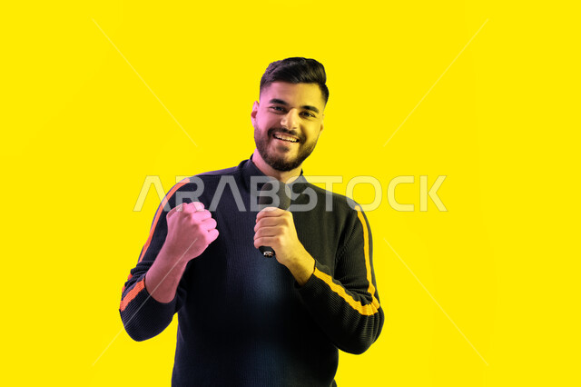 ايماءات تدل على الاندماج والتفاعل مع الغناء، الاستمتاع بأداء المواهب الصوتية، بورتريه مقرب لرجل عربي اماراتي خليجي يرتدي الزي الكاجوال يمسك بيده المايكروفون و يغني بحماس، شاب سعودي يقوم بممارسة موهبة الغناء، تسجيل مقطع صوتي غنائي، خلفية صفراء