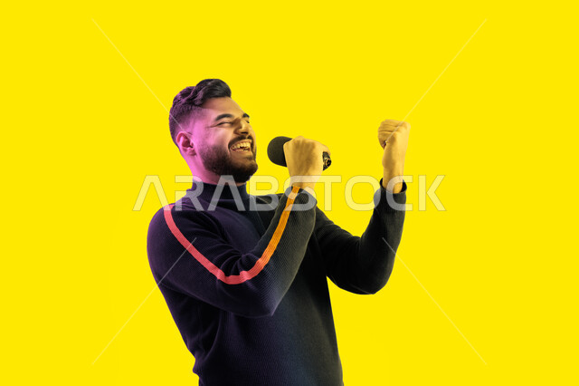 ايماءات تدل على الاندماج والتفاعل مع الغناء، الاستمتاع بأداء المواهب الصوتية، بورتريه مقرب لرجل عربي اماراتي خليجي يرتدي الزي الكاجوال يمسك بيده المايكروفون و يغني بحماس، شاب سعودي يقوم بممارسة موهبة الغناء، تسجيل مقطع صوتي غنائي، خلفية صفراء