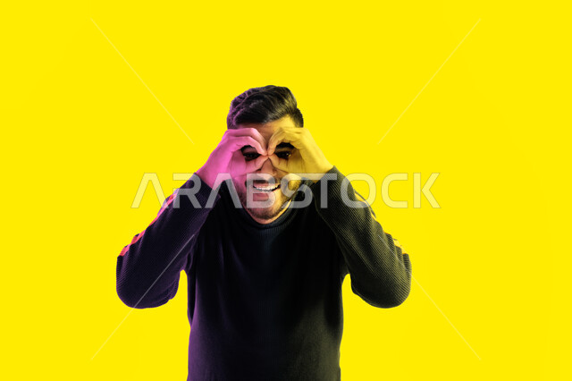 تعبيرات وجه تدل على الفرح والسعادة لرؤية شيء ما، بورتريه مقرب لشاب سعودي يضع يده على عينيه كالناظور، الاستكشاف و المغامرة مع فريق الكشافة، رجل إماراتي عربي خليجي مستكشف يرتدي الزي الكاجوال، البحث والنظرة المستقبلية، خلفية صفراء