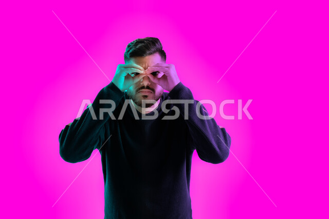 تعبيرات وجه تدل التركيز والبحث لرؤية شيء ما، بورتريه مقرب لشاب سعودي يضع يده على عينيه كالناظور، الاستكشاف و المغامرة مع فريق الكشافة، رجل إماراتي عربي خليجي مستكشف يرتدي الزي الكاجوال، البحث والنظرة المستقبلية، خلفية ملونة