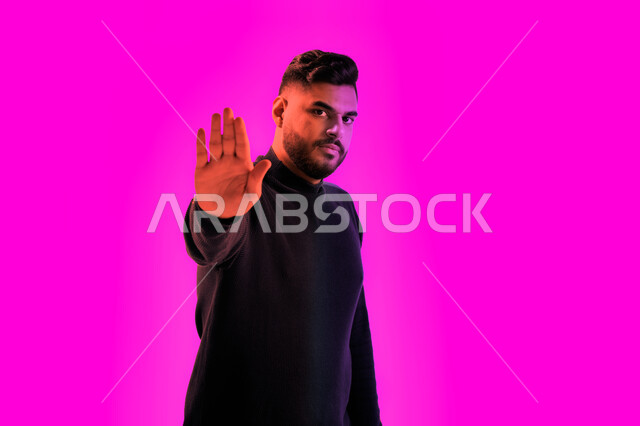 الشعور بالخوف و الإنزعاج, بورتريه مقرب لشاب سعودي يقوم بجلسة تصوير يرتدي الزي الكاجوال، إيماءات وجه غاضبة ويدين تدل على المنع والتوقف عن فعل شيء ما، رجل عربي خليجي إماراتي يعبر عن الرفض والإمتناع وايقاف أمر ما، خلفية ملونة