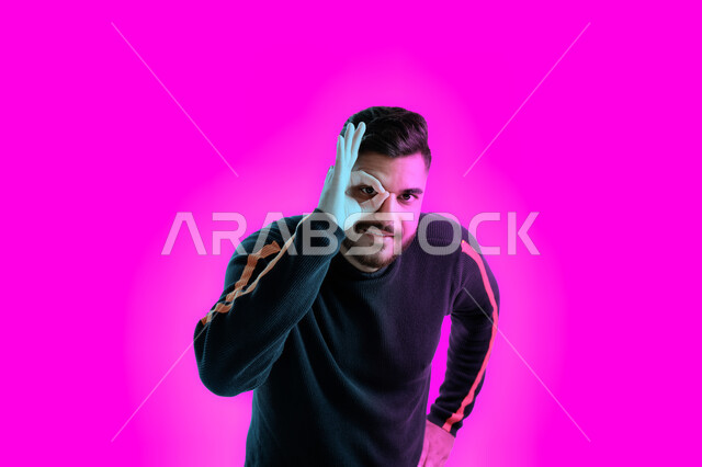 الشعور بالسعادة من أجل إكتشاف الحقائق، بورتريه مقرب لشاب عربي خليجي اماراتي يضع يده على عينه للتدقيق والنظر الى شيئ محدد، شاب سعودي يرتدي الزي الكاجوال، إشارات يد تدل على التركيز على شيء ما، خلفية ملونة