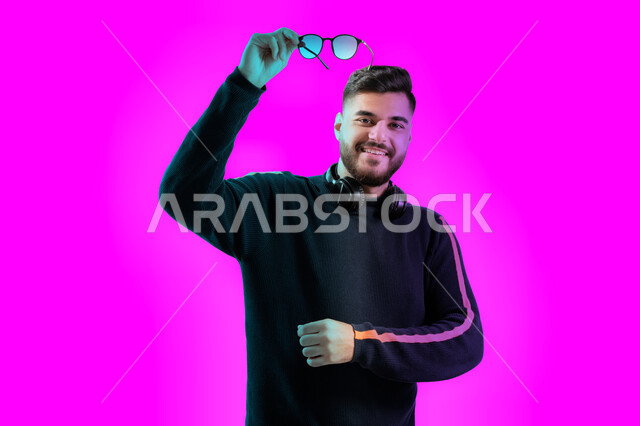 الشعور بالسعادة والفرح لإمتلاك نظارة شمسية جديدة، مواهب غنائية فنية، رجل عربي اماراتي عربي خليجي يضع الهيدفون على رقبته، إيماءات و تعابير وجه تدل على السعادة، بورتريه مقرب لشاب سعودي يرتدي الزي الكاجوال يرفع النظارات الشمسيه للأعلى، خلفية ملونة