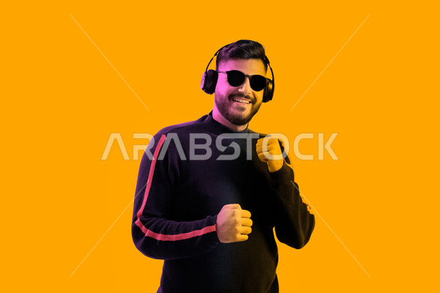 إيماءات و تعابير وجه ويدين تدل على الاندماج مع الأغنية، الاستماع إلى الأغاني بواسطة الهيدفون، بورتريه مقرب لشاب سعودي يرتدي النظارات الشمسية وسماعات رأس في استديو صوتي، رجل عربي إماراتي خليجي يرتدي الزي الكاجوال ويبتسم، خلفية ملونة