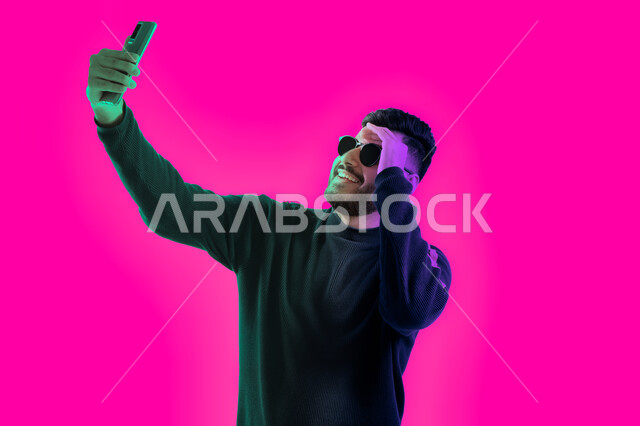 Taking memorial photos with different gestures using the mobile phone, answering a phone video call, a close-up portrait of a Saudi young man wearing casual clothes and sunglasses holding a mobile phone in his hand, an Arab Emirati Gulf man conducting a l