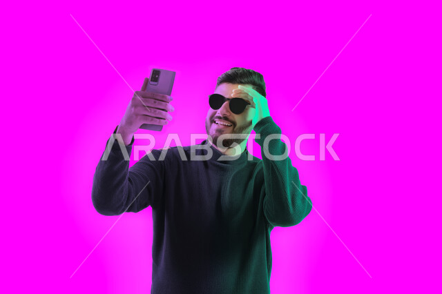 إجراء بث مباشر على إحدى وسائل التواصل الاجتماعي, التقاط صور تذكارية بإيماءات مختلفة باستخدام الجوال، بوتريه مقرب لشاب سعودي يرتدي الزي الكاجوال و النظارات الشمسية يحمل بيده هاتف محمول, رجل عربي اماراتي خليجي يقوم بالرد على مكالمة فيديو هاتفية, خلفية ملونة