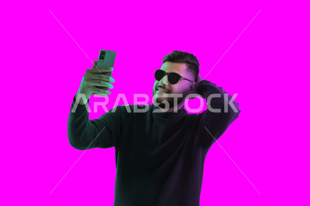 إجراء بث مباشر على إحدى وسائل التواصل الاجتماعي, التقاط صور تذكارية بإيماءات مختلفة باستخدام الجوال، بوتريه مقرب لشاب سعودي يرتدي الزي الكاجوال و النظارات الشمسية يحمل بيده هاتف محمول, رجل عربي اماراتي خليجي يقوم بالرد على مكالمة فيديو هاتفية, خلفية ملونة