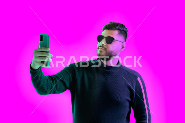 الرد على مكالمة فيديو هاتفية, التقاط صور تذكارية بإيماءات مختلفة باستخدام الجوال، بوتريه مقرب لشاب سعودي يرتدي الزي الكاجوال و النظارات الشمسية يحمل بيده هاتف محمول, رجل عربي اماراتي خليجي يقوم بإجراء بث مباشر على وسائل التواصل الاجتماعي، خلفية ملونة.