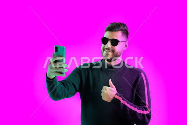Answering a phone video call, taking souvenir photos with admiration gestures using a mobile phone, a close-up portrait of a Saudi young man wearing casual clothes and sunglasses holding a mobile phone in his hand, an Arab Emirati Gulf man conducting a li