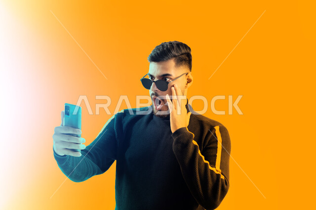 ايماءات دهشة و استغراب, التقاط صور تذكارية بإيماءات مختلفة باستخدام الجوال، بوتريه مقرب لشاب سعودي يرتدي الزي الكاجوال و النظارات الشمسية يحمل بيده هاتف محمول, رجل عربي اماراتي خليجي يقوم بالرد على مكالمة فيديو هاتفية, خلفية صفراء.
