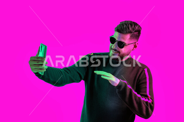 Taking memorial photos with different gestures using the mobile phone, a close-up portrait of a Saudi young man wearing casual clothes and sunglasses holding a mobile phone in his hand, an Arab Emirati Gulf man conducting a live broadcast on social media,