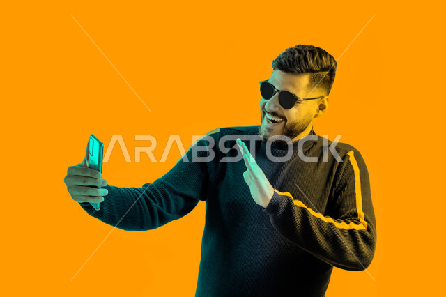 التقاط صور تذكارية بإيماءات مختلفة باستخدام الجوال، بوتريه مقرب لشاب سعودي يرتدي الزي الكاجوال و النظارات الشمسية يحمل بيده هاتف محمول, رجل عربي اماراتي خليجي يقوم بإجراء بث مباشر على وسائل التواصل الاجتماعي، الرد على مكالمة فيديو هاتفية, خلفية صفراء.