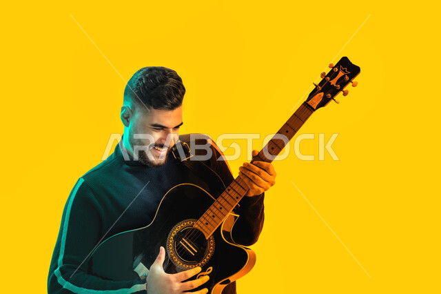 العزف على آلة وترية، بورتريه مقرب لشاب سعودي يقوم بالعزف على الجيتار, شاب عربي اماراتي خليجي يرتدي الزي الكاجوال يعزف على إحدى الآلات الموسيقية, ايماءات وجه و يدين تدل على الحماس و الشغف, مجال صوتي و فني طربي, خلفية صفراء
