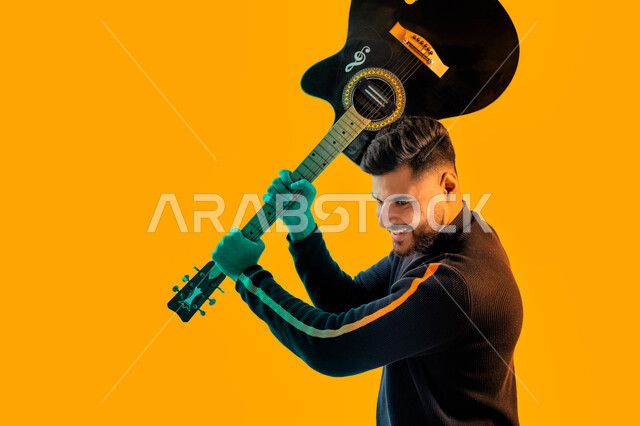 ايماءات وجه و يدين تدل على الحماس و الشغف, العزف على آلة وترية، بورتريه مقرب لشاب سعودي عربي خليجي مبتسم يقوم برفع الجيتار للأعلى, شاب اماراتي يرتدي الزي الكاجوال يحمل بيديه إحدى الآلات الموسيقية, مجال صوتي و فني طربي, خلفية صفراء