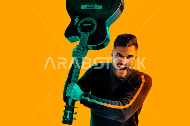 مجال صوتي و فني طربي, ايماءات وجه و يدين تدل على الحماس و الشغف, العزف على آلة وترية، بورتريه مقرب لشاب سعودي يقوم برفع الجيتار للأعلى, شاب اماراتي عربي خليجي يرتدي الزي الكاجوال يحمل بيديه إحدى الآلات الموسيقية, خلفية صفراء