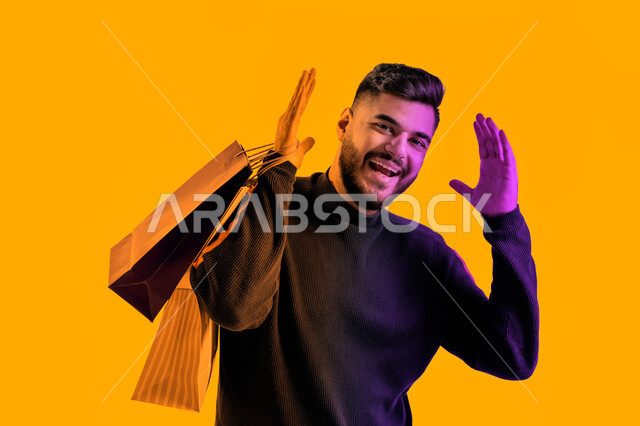 ايماءات وجه و يدين تدل على السعادة و المتعة بإقتناء مستلزمات جديدة, الاستمتاع بالشراء والتبضع، بورتريه مقرب لشاب سعودي يرتدي الزي الكاجوال يحمل بيده أكياس التسوق، رجل عربي اماراتي خليجي يقوم بالتسوق من المتاجر والأسواق, خلفيه صفراء. 
