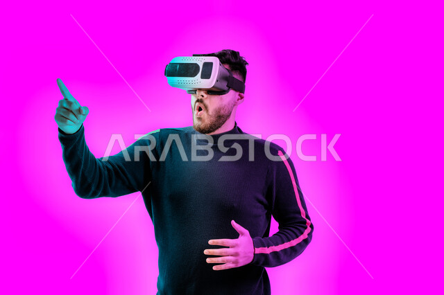 التفاعل ومحاكاة العالم الخيالي، رجل اماراتي يقوم بمشاهدة أفلام عبر نظارات الواقع الإفتراضي يشير بإصبعه لشيء ما، ايماءات تدل على الدهشة والاستغراب, بورتريه مقرب لشاب عربي خليجي سعودي يرتدي الزي الكاجوال ونظارة الواقع الافتراضي ثلاثية الأبعاد, خلفية ملونة