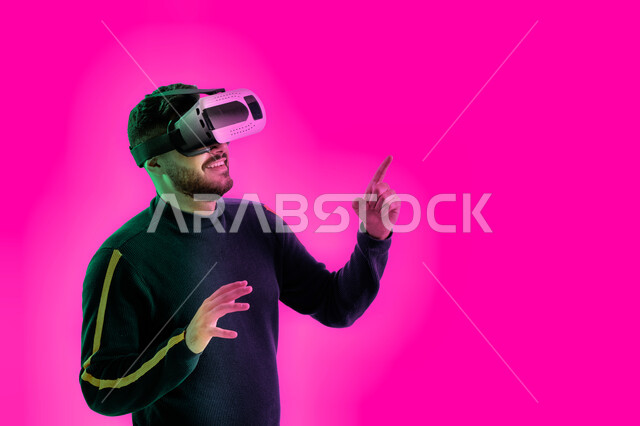 محاكاة العالم الخيالي والتفاعل مع الفيلم، شاب عربي خليجي اماراتي يقوم بمشاهدة أفلام عبر نظارات الواقع الإفتراضي يشير بإصبعه لشيء ما، ايماءات تدل على المتعة, بورتريه مقرب لشاب سعودي يرتدي الزي الكاجوال ونظارة الواقع الافتراضي ثلاثية الأبعاد, خلفية ملونة