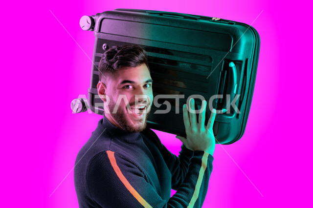 إيماءات وجه تدل على الحماس للقيام برحلة سياحية, الاستعداد و تجهيز الأمتعة للسفر في جولة سياحية، بورتريه مقرب لرجل سعودي مبتسم يحمل حقيبة سفر سوداء، شاب عربي اماراتي خليجي يرتدي الزي الكاجوال ينظر الى الكاميرا, خلفية ملونة