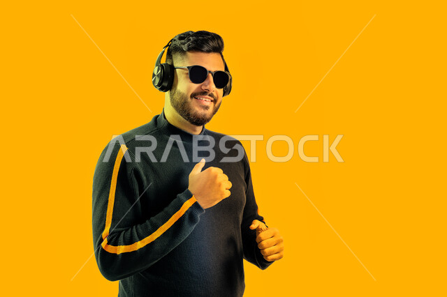 إيماءات وجه تدل على الاندماج مع الأغنية، بورتريه مقرب لشاب سعودي يرتدي  النظارات الشمسية وسماعة رأس في استديو صوتي, رجل عربي اماراتي خليجي يرتدي الزي الكاجوال يقوم بالإستماع للأغاني بإستخدام الهيدفون، مواهب غنائية فنية, خلفية صفراء