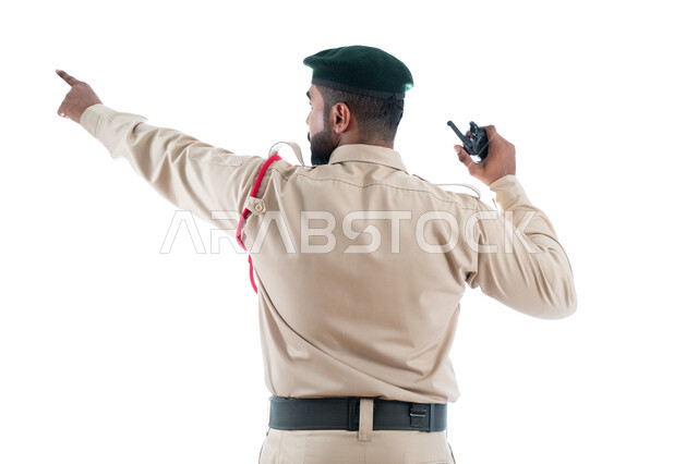 استخدام جهاز الإرسال والإستقبال اللاسلكي، تعزيز الثقة بالشرطة، بورتريه من الخلف لشرطي عربي خليجي إماراتي يرتدي الزي العسكري ويشير بإصبع السبابة نحو شيء ما، حركة إصبع وإشارات، الحفاظ الأمن والأمان والطمأنينة، تأمين المنشأت وحماية الأفراد، خلفية بيضاء