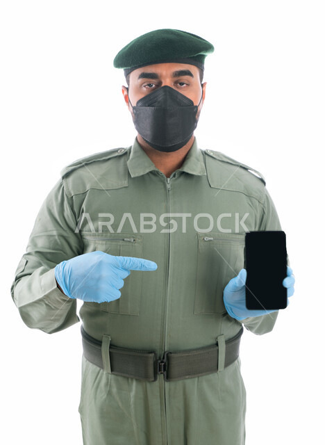إستخدام تطبيقات الأمن والسلامهة في الهاتف الذكي، بورتريه لمحقق أدلة جنائية عربي خليجي إماراتي يرتدي الكمامة الواقية والقفازات وبدلة الأمن الخضراء، الإشارة بإصبع السبابة نحو الجوال بشاشة سوداء فارغة، برامج وانظمة حماية الهاتف المحمول، خلفية بيضاء