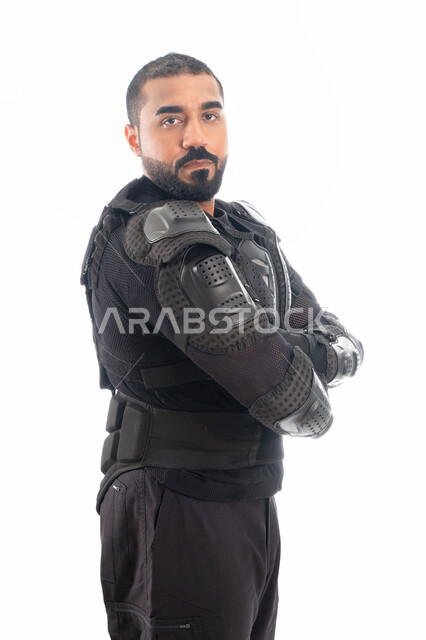 الوقوف بإستقامة وإنضباط، بورتريه لشرطي عربي خليجي إماراتي من قوات التدخل السريع (سوات) يرتدي البدلة العسكرية السوداء بأيدي متقاطعة، الحفاظ على الأمن والأمان، إيماءات وجه ويدين تدل على الثقة، خلفية بيضاء
