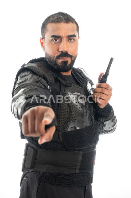 حركة اصبع واشارات، بورتريه لشرطي عربي خليجي إماراتي من قوات التدخل السريع (سوات) يرتدي البدلة العسكرية السوداء ويشير باصبع السبابة نحو شيء ما، استخدام جهاز الارسال والاستقبال اللاسلكي، الحفاظ على الأمن والأمان، خلفية بيضاء