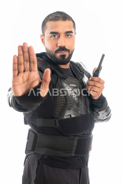استخدام جهاز الارسال والاستقبال اللاسلكي، بورتريه لشرطي عربي خليجي إماراتي يرتدي البدلة العسكرية السوداء ويشير بإشارة قف، قوات التدخل السريع (سوات)، الحفاظ على الأمن والأمان، إيماءات يدين تدل على التوقف والإنتظار، حركة يد وإشارات، خلفية بيضاء
