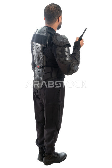 صورة الجسم بالطول الكامل، بورتريه من الخلف لشرطي عربي خليجي إماراتي من قوات التدخل السريع (سوات) يرتدي البدلة العسكرية السوداء، استخدام جهاز الإرسال والإستقبال اللاسلكي، الحفاظ على الأمن والأمان، الوقوف باستعداد وانضباط، خلفية بيضاء