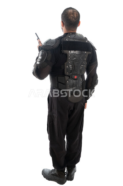 استخدام جهاز الإرسال والإستقبال اللاسلكي، بورتريه من الخلف لشرطي عربي خليجي إماراتي من قوات التدخل السريع (سوات) يرتدي البدلة العسكرية السوداء، الحفاظ على الأمن والأمان، الوقوف باستعداد وانضباط، صورة الجسم بالطول الكامل، خلفية بيضاء