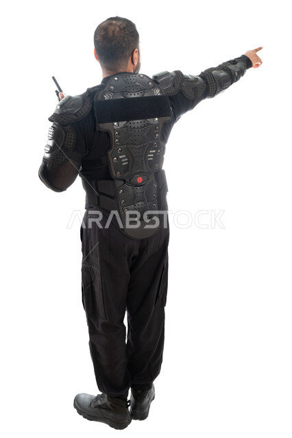 الحفاظ على الأمن والأمان، بورتريه من الخلف لشرطي عربي خليجي إماراتي من قوات التدخل السريع (سوات) يرتدي البدلة العسكرية السوداء ويستخدم جهاز الإرسال والإستقبال اللاسلكي ويشير باصبع السبابة، الإشارة نحو شيء ما، صورة الجسم بالطول الكامل، خلفية بيضاء