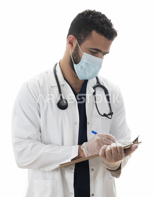 Preparing medical reports, a portrait of a Saudi doctor who records the results of medical examinations in the medical record, an Arab Emirati Gulf youth wearing a medical coat and a muzzle and efficiently providing treatment services to patients, working