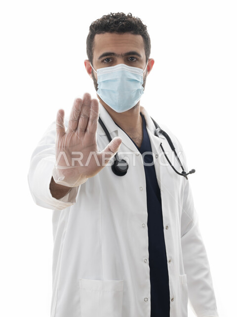العمل في القطاع الصحي، بورتريه مقرب لطبيب سعودي يرتدي القفازات و المعطف الطبي والكمامة لاتباع توصيات وزارة الصحة للحفاظ على السلامة , شاب عربي اماراتي خليجي يشير بيده بإيماءات تدل على تجنب انتشار العدوى، مهن ووظائف طبية في القطاع الصحي