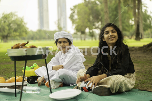 مفهوم العلاقات الودية بين الاخوه، انشطة صيفيه ترفيهيه, ولد عربي اماراتي خليجي يرتدي  الكندورة و العمامة البيضاء في إحدى المنتزهات مع شقيقته في الخارج، ولد وبنت سعوديان في الخارج يستمتعان في التنزه في الهواء الطلق في الحديقه