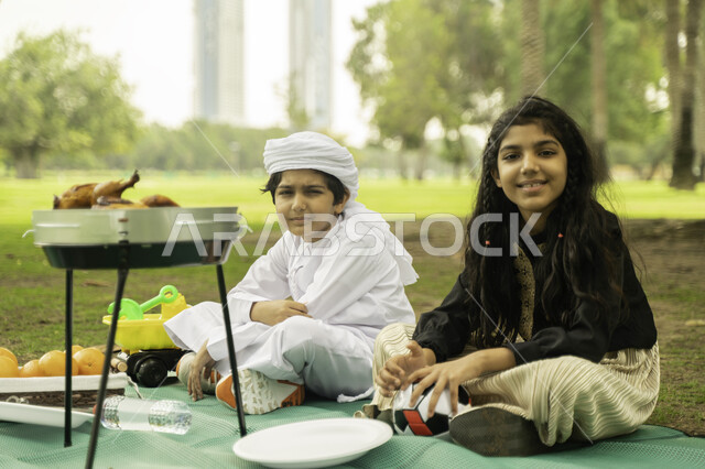 النظر الى الكاميرا, مفهوم العلاقات الودية بين الاخوه، انشطة صيفيه ترفيهيه, ولد عربي اماراتي خليجي يرتدي  الكندورة و العمامة البيضاء في إحدى المنتزهات مع شقيقته في الخارج، ولد وبنت سعوديان في الخارج يستمتعان في التنزه في الهواء الطلق في الحديقه