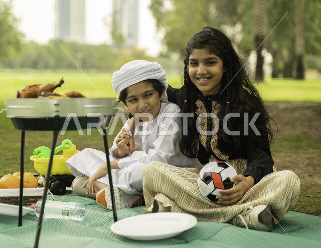 ايماءات تدل على الفرح، النظر الى الكاميرا, مفهوم التعاطف والحب بين الاخوه، انشطة صيفيه ترفيهيه, صورة لبنت عربية اماراتية خليجية تقوم بضم شقيقها يرتدي الكندورة و العمامة البيضاء في إحدى المزارع، ولد وبنت سعوديان في الخارج يستمتعان في التنزه في الهواء الطلق